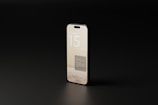 A modern smartphone displayed at an angle on a dark surface with a minimalistic interface. The text on the screen reads 'iPhone 15 mockups.' The phone has a sleek, reflective surface with a muted color scheme that accentuates its smooth design.