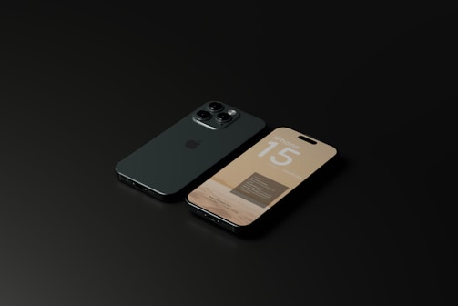 A pair of modern smartphones placed side by side on a dark surface. One of the phones is showing its display screen with the text 'iPhone 15,' while the other phone’s back is visible, featuring a triple-camera setup.