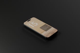 A smartphone with a sleek design is lying on a dark surface. The screen displays information with the text 'iPhone 15' prominently featured, along with some unreadable smaller text.