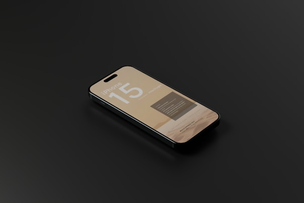 A smartphone with a sleek design is lying on a dark surface. The screen displays information with the text 'iPhone 15' prominently featured, along with some unreadable smaller text.