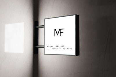 A modern square sign with the letters 'MF' is mounted on a wall. The sign has a minimalistic design with a black frame and a white background. The text 'MOCKUPFREE.NET' and 'REALISTIC MOCKUPS' is written at the bottom in smaller font. The background is a smooth, light-colored wall with subtle shadows.