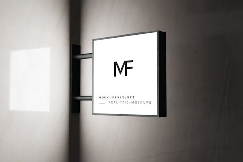 A modern square sign with the letters 'MF' is mounted on a wall. The sign has a minimalistic design with a black frame and a white background. The text 'MOCKUPFREE.NET' and 'REALISTIC MOCKUPS' is written at the bottom in smaller font. The background is a smooth, light-colored wall with subtle shadows.