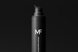 A sleek, black bottle with a pump dispenser top. The bottle has a matte finish and features the text 'MF' in large white letters, with 'pump bottle mockups' written vertically beneath it. The background is also black, creating a minimalist and modern aesthetic.