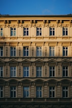 A detailed architectural facade featuring multiple rows of windows adorned with ornate moldings and decorative embellishments. The building displays classic European design elements, with a symmetrical arrangement and intricate stone carvings. The warm sunlight casts shadows across the facade, enhancing its texture and depth.