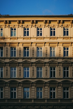 A detailed architectural facade featuring multiple rows of windows adorned with ornate moldings and decorative embellishments. The building displays classic European design elements, with a symmetrical arrangement and intricate stone carvings. The warm sunlight casts shadows across the facade, enhancing its texture and depth.