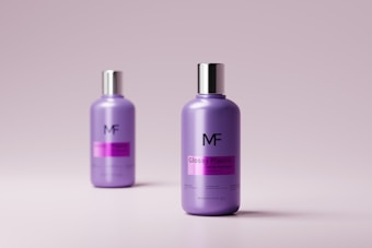Two purple bottles with silver caps are placed against a light pink background. The bottle in the foreground is more prominent, displaying the label 'Glossy Plastic' with the MF logo. The second bottle is positioned slightly blurred, emphasizing the leading bottle.