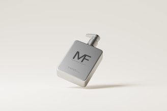 A sleek, metallic dispenser bottle with a pump, featuring the letters 'MF' on the front. The bottle appears to be floating against a minimalistic, light beige background, emphasizing its modern design.