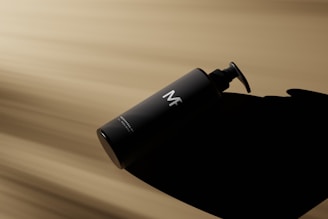 A black cylindrical bottle with a pump dispenser lying on its side against a blurred beige and brown background. The bottle casts a dark shadow and has the letters 'MF' prominently displayed in white on its front. The lighting emphasizes the sleek and minimalist design of the bottle.