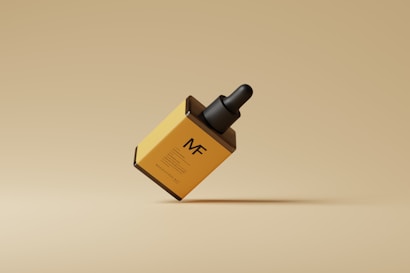 A minimalist image features a yellow rectangular box tilted slightly, with a black dropper cap on top. The box has the letters 'MF' printed prominently on the front, with additional smaller text below. It appears to be a cosmetic or skincare product placed against a beige background.