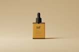 An amber-colored glass dropper bottle with a black cap, labeled with the letters 'MF', is centered against a beige background. The minimalist design emphasizes a sophisticated and elegant appearance.
