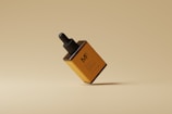 A minimalistic still life of a brown and orange dropper bottle with a black cap. The bottle appears to be floating against a monochromatic beige background, creating a clean and modern aesthetic.