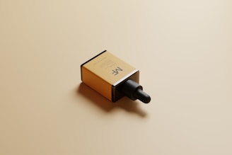 A rectangular cardboard box with a minimalist design lies on a light tan surface. The box prominently features a dropper cap and has the initials 'MF' printed on its side, indicating it might contain a cosmetic or skincare product. The lighting casts soft shadows, adding a subtle dimension to the scene.