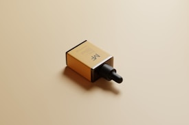 A rectangular cardboard box with a minimalist design lies on a light tan surface. The box prominently features a dropper cap and has the initials 'MF' printed on its side, indicating it might contain a cosmetic or skincare product. The lighting casts soft shadows, adding a subtle dimension to the scene.