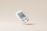 A digital glucose meter displaying a reading of 106 mg/dL. The device is predominantly white with purple buttons labeled with 'M' and 'S'. A test strip is inserted into the bottom of the meter. The background is a plain, light beige color.