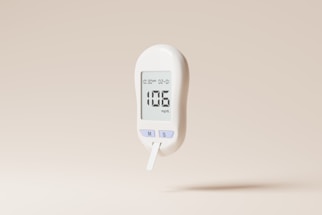 A white digital glucose meter displaying a reading of 106 mg/dL is set against a plain beige background. The device includes buttons labeled 'M' and 'S' and shows a timestamp on its screen.
