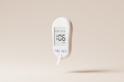 A white digital glucose meter displaying a reading of 106 mg/dL is set against a plain beige background. The device includes buttons labeled 'M' and 'S' and shows a timestamp on its screen.