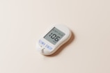 A white digital glucose meter with a display showing a reading of 106 mg/dL is placed on a light beige surface. The device includes buttons labeled 'M', 'S', and a test strip protruding from the bottom.