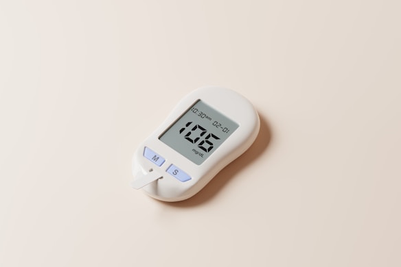 A white digital glucose meter with a display showing a reading of 106 mg/dL is placed on a light beige surface. The device includes buttons labeled 'M', 'S', and a test strip protruding from the bottom.