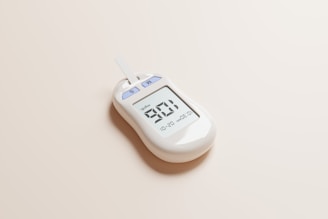 A digital glucometer displaying a reading of 98 mg/dL, placed on a neutral light-colored surface. The device features a test strip inserted at the top and two buttons labeled 'S' and 'M'.
