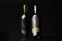 Two elegant wine bottles with dark labels and gold caps stand against a black background. The lighting highlights the bottles, creating a sophisticated and luxurious feel.
