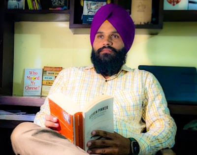 A man with a beard wearing a purple turban and a plaid shirt sits in a cozy setting, deeply engrossed in reading a book titled 'Steven Pinker'. Behind him, a wooden shelf holds various books, including 'Who Moved My Cheese?' and 'How to Read a Book'. The background is softly lit, creating a serene and intellectually stimulating atmosphere.
