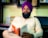 A man with a beard wearing a purple turban and a plaid shirt sits in a cozy setting, deeply engrossed in reading a book titled 'Steven Pinker'. Behind him, a wooden shelf holds various books, including 'Who Moved My Cheese?' and 'How to Read a Book'. The background is softly lit, creating a serene and intellectually stimulating atmosphere.