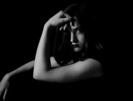 A black and white portrait features a person with a contemplative expression, having their hand resting on their face and slightly covering one eye. The lighting is dramatic, creating striking contrasts and emphasizing the person's facial features against a dark background.