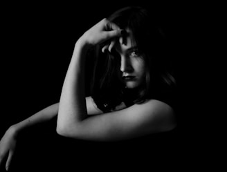A black and white portrait features a person with a contemplative expression, having their hand resting on their face and slightly covering one eye. The lighting is dramatic, creating striking contrasts and emphasizing the person's facial features against a dark background.