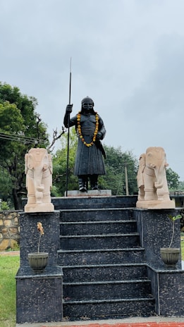 A statue of a warrior standing on a multi-step platform is flanked by two smaller elephant statues. The warrior statue is adorned with a garland and holds a spear. The setting is outdoors with green trees and cloudy skies in the background.