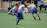 A group of young children are actively playing soccer on a green sports field. They are wearing blue and gray soccer uniforms. One child is dribbling a soccer ball while others are running around. Spectators can be seen sitting in the background near a fence, watching the match.