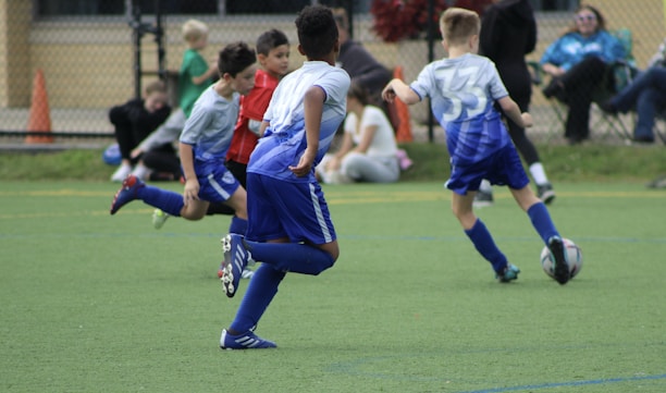 A group of young children are actively playing soccer on a green sports field. They are wearing blue and gray soccer uniforms. One child is dribbling a soccer ball while others are running around. Spectators can be seen sitting in the background near a fence, watching the match.
