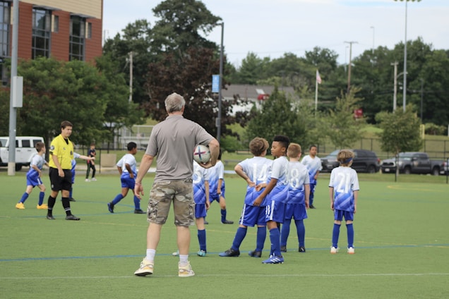Several young boys in blue and white soccer jerseys gather on a green field with a coach holding a soccer ball. A referee in a yellow shirt is also present. Trees, a building, and parked cars are visible in the background. The boys appear to be listening to instructions from the coach.