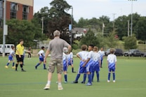 Several young boys in blue and white soccer jerseys gather on a green field with a coach holding a soccer ball. A referee in a yellow shirt is also present. Trees, a building, and parked cars are visible in the background. The boys appear to be listening to instructions from the coach.