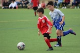 Two young boys are in soccer uniforms, actively engaged in a match on a grassy field. One boy in a red jersey is dribbling a soccer ball, while the other in a blue and gray jersey is closely following him, seemingly trying to defend or intercept the ball. In the background, other players and spectators can be seen, blurred to emphasize the action in the foreground.