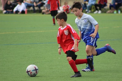 Two young boys are in soccer uniforms, actively engaged in a match on a grassy field. One boy in a red jersey is dribbling a soccer ball, while the other in a blue and gray jersey is closely following him, seemingly trying to defend or intercept the ball. In the background, other players and spectators can be seen, blurred to emphasize the action in the foreground.