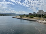 A scenic riverside view with a wide expanse of water in the foreground and a grassy park area with trees along the shore. In the background, there are modern buildings, likely part of an urban environment, with hills visible in the distance under a partly cloudy sky.