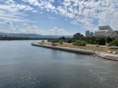 A scenic riverside view with a wide expanse of water in the foreground and a grassy park area with trees along the shore. In the background, there are modern buildings, likely part of an urban environment, with hills visible in the distance under a partly cloudy sky.
