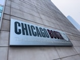 A large rectangular sign attached to a concrete building displays the text 'CHICAGO BOOTH' in bold letters, with 'The University of Chicago Booth School of Business' subtitled beneath it. Part of a city skyline is visible in the background.