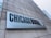 A large rectangular sign attached to a concrete building displays the text 'CHICAGO BOOTH' in bold letters, with 'The University of Chicago Booth School of Business' subtitled beneath it. Part of a city skyline is visible in the background.
