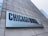 A large rectangular sign attached to a concrete building displays the text 'CHICAGO BOOTH' in bold letters, with 'The University of Chicago Booth School of Business' subtitled beneath it. Part of a city skyline is visible in the background.