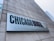 A large rectangular sign attached to a concrete building displays the text 'CHICAGO BOOTH' in bold letters, with 'The University of Chicago Booth School of Business' subtitled beneath it. Part of a city skyline is visible in the background.