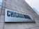 A large rectangular sign attached to a concrete building displays the text 'CHICAGO BOOTH' in bold letters, with 'The University of Chicago Booth School of Business' subtitled beneath it. Part of a city skyline is visible in the background.