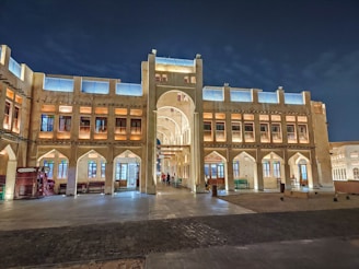 A traditional Middle Eastern marketplace building with arched windows and decorative lighting, creating a warm and inviting atmosphere. The structure features multiple floors with intricate architectural details, including a large archway leading to an interior courtyard. The setting appears to be during the evening with lights illuminating the facade.