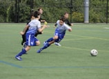 A group of young boys are actively playing soccer on a green sports field. One boy in a blue and white uniform is lunging forward, closely followed by three others in darker uniforms. The ball is in motion on the field, indicating an intense play.
