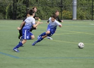 A group of young boys are actively playing soccer on a green sports field. One boy in a blue and white uniform is lunging forward, closely followed by three others in darker uniforms. The ball is in motion on the field, indicating an intense play.
