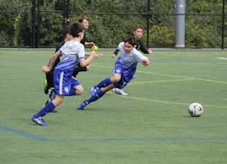 A group of young boys are actively playing soccer on a green sports field. One boy in a blue and white uniform is lunging forward, closely followed by three others in darker uniforms. The ball is in motion on the field, indicating an intense play.