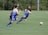 A group of young boys are actively playing soccer on a green sports field. One boy in a blue and white uniform is lunging forward, closely followed by three others in darker uniforms. The ball is in motion on the field, indicating an intense play.