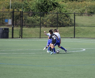 A group of young soccer players is engaged in a game on a grassy field. One player in a black jersey with the number 23 is actively dribbling the ball while two players in blue jerseys attempt to contest. The background features a chain-link fence and lush green trees.