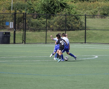 A group of young soccer players is engaged in a game on a grassy field. One player in a black jersey with the number 23 is actively dribbling the ball while two players in blue jerseys attempt to contest. The background features a chain-link fence and lush green trees.
