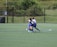 A group of young soccer players is engaged in a game on a grassy field. One player in a black jersey with the number 23 is actively dribbling the ball while two players in blue jerseys attempt to contest. The background features a chain-link fence and lush green trees.